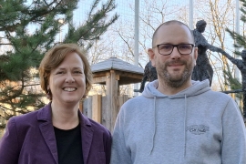 Eine Frau in Lila Blaizer mit roten Haaren und ein Mann mit grauem Hoodie, Bart und sehr kurzen Haaren lächeln vor einer Don Bosco Skulptur in die Kamera.