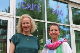 Martina Köhne, blonde, schulterlange Haare, dunkelgrünes Obertteil, und Isolde Gertig, kurze dunkle Haare, buntes Halstuch, weißes Shirt, lächeln in die Kamera. Sie stehen vor dem Café MiA, im Hintergrund sind die Scheiben und der darauf angebrachte Schriftzug mit dem Namen des Cafés zu sehen.