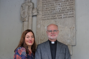 Julia Dahmen und P. Alfons Friedrich stehen vor einer Tafel, die die Namen der bisherigen Pfarrer der Pfarrei St. Wolfgang aufführt.