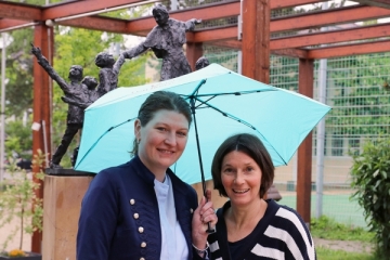 Zwei Frauen stehen lächelnd unter einem hellblauen Regenschirm vor einer Statue von Don Bosco.